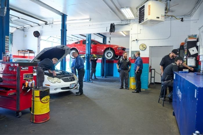 Several workers repair cars and talk inside a well-lit garage, with tools and vehicles in the background.