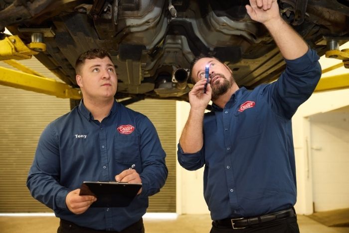 Mechanics examine the undercarriage of a raised car, one shining a light while the other takes notes on a clipboard.