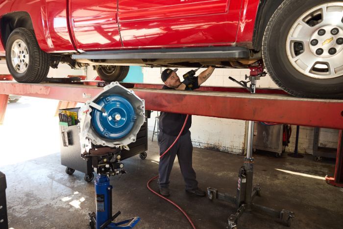 Technician repairs the underside of a red truck on a lift in a garage, with a blue transmission unit in the foreground.