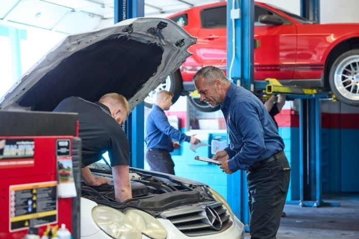 Mechanics work on a car engine in a garage while another red car is elevated on a hydraulic lift in the background.