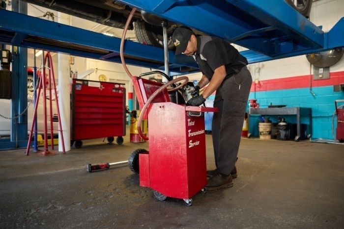 A mechanic in uniform works with a red transmission service machine in an auto repair shop under a car lift.