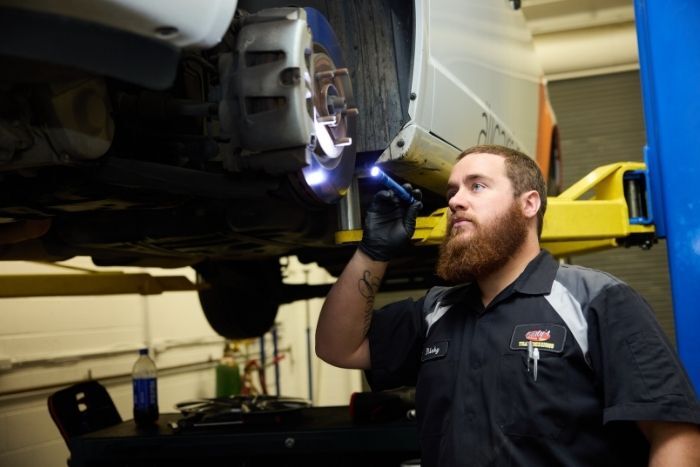 Mechanic inspects the underside of a vehicle on a lift, holding a flashlight and wearing a uniform in an auto shop.